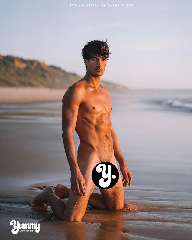 Model Romain Beziau kneeling on a wet sandy shoreline by the ocean during sunset in an editorial beach shoot.