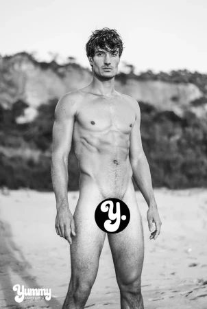 Black and white full-length portrait of Romain Beziau standing on a sandy beach against a soft-focus coastal cliff background.