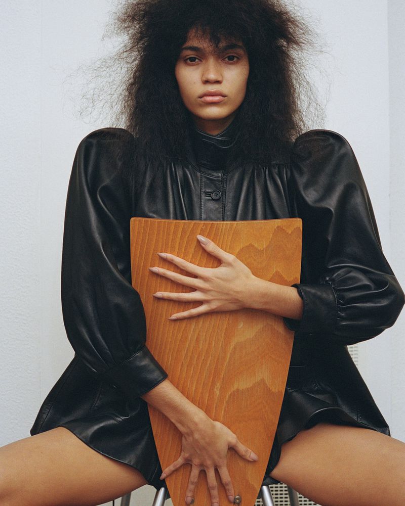 A young woman with natural hair poses in a studio wearing a black leather outfit while sitting on a stool and holding a wooden object.