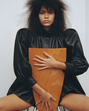 A young woman with natural hair poses in a studio wearing a black leather outfit while sitting on a stool and holding a wooden object.