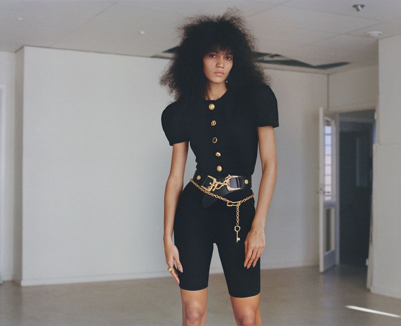 A slender woman with curly hair poses in a black top and shorts with a gold chain belt in a sparsely decorated interior setting.