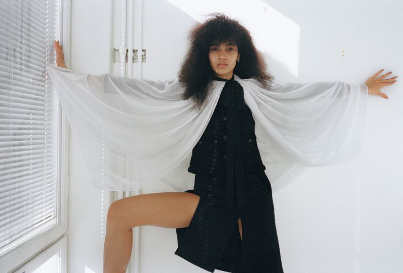 A woman with afro-style hair wearing a sheer white cape and black outfit, posing in front of a window with white blinds.