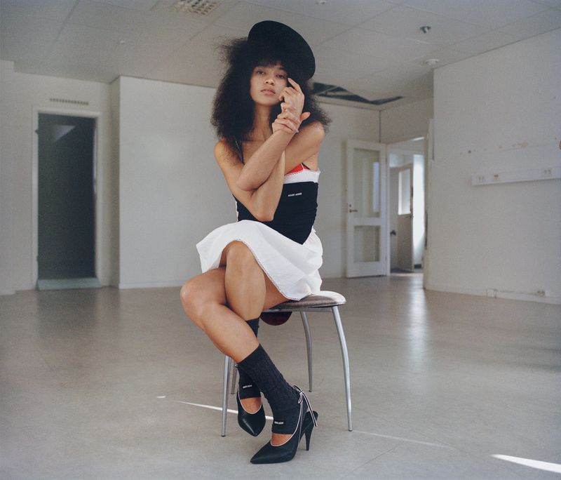 Woman with afro and black hat sitting on metal stool in an empty room wearing a black and white dress, socks, and pointed toe heels.