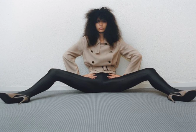 Woman with afro hairstyle poses sitting on the floor wearing a trench coat, black leggings and beige high heels against a white wall.