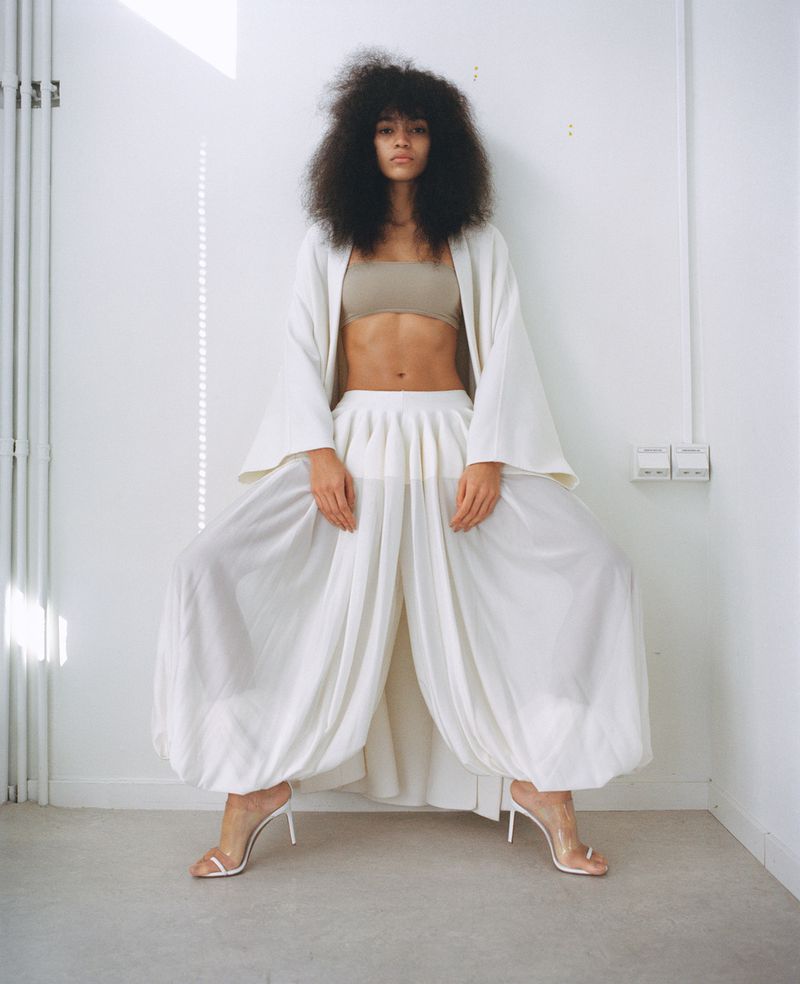 A woman with an afro poses in wide-leg pants and a beige top indoors, complemented by white heels and a flowing white jacket.
