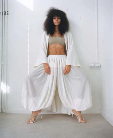 A woman with an afro poses in wide-leg pants and a beige top indoors, complemented by white heels and a flowing white jacket.