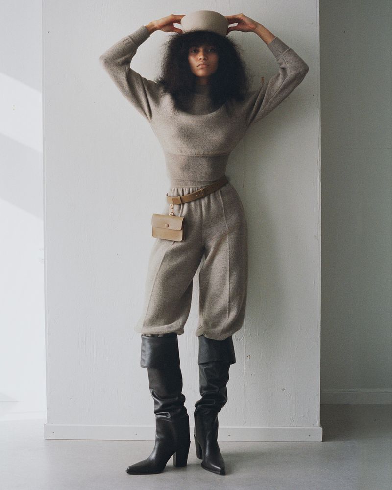 A woman in a light brown sweater, cropped pants, and black boots poses against a white wall in a fashion editorial photograph.