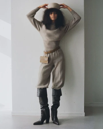 A woman in a light brown sweater, cropped pants, and black boots poses against a white wall in a fashion editorial photograph.
