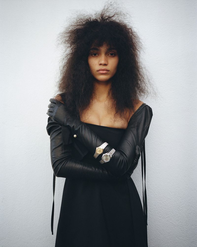 A woman with dark, voluminous hair wearing a black dress and long leather gloves with two wristwatches posing in a studio.