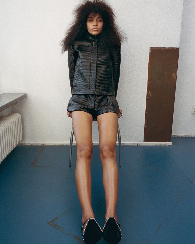 An androgynous fashion model with dark curly hair is sitting on a metal stool in a photo studio wearing a black textured matching short set.