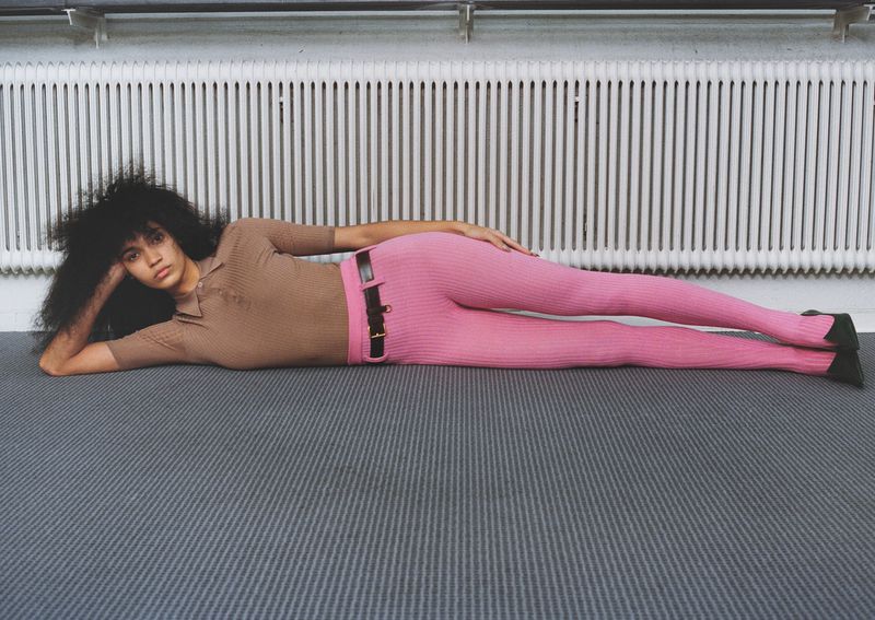 Woman with curly hair poses while lying on a gray patterned carpet wearing a brown top and pink ribbed leggings in an indoor setting.