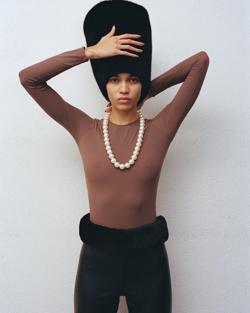 Model Raynara Negrine wearing an oversized black fur hat, brown long-sleeve top, and chunky pearl necklace against a white background.