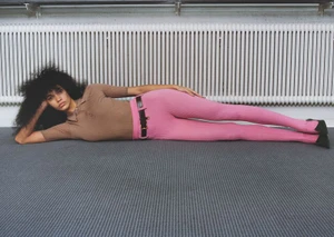 Model Raynara Negrine reclines in a brown polo shirt and bright pink leggings on a carpeted floor in front of a white radiator.