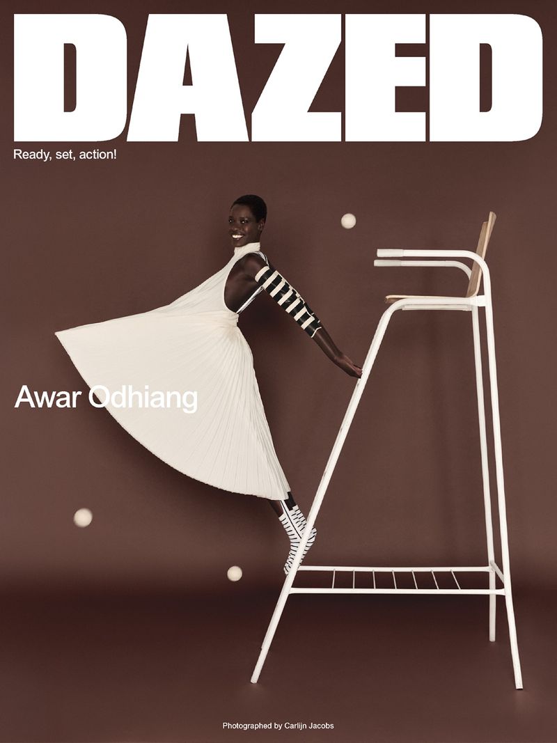 Model Awar Odhiang in a white pleated dress posing on a white umpire chair against a brown studio backdrop for Dazed Magazine.