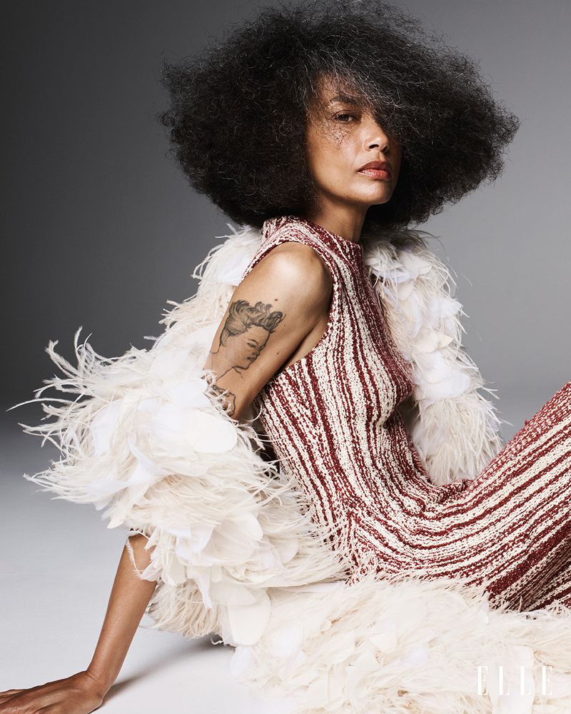 Model Stephanie Cavalli seated in a striped knit dress and voluminous white feathered wrap against a neutral studio backdrop.
