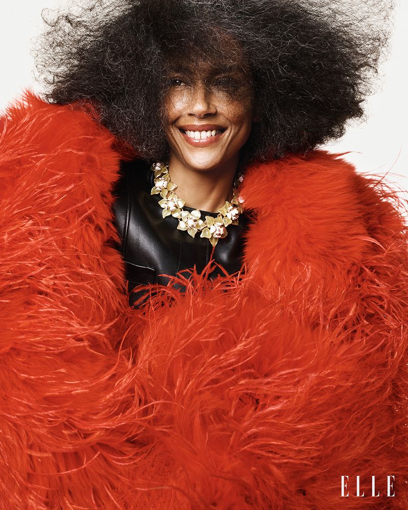 Stephanie Cavalli smiling in a voluminous red feather coat and floral collar necklace against a plain white studio background.