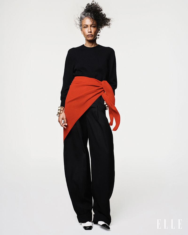 Model Stephanie Cavalli posing in a black crewneck sweater, wide-leg trousers, and an orange waist wrap against a white studio background.