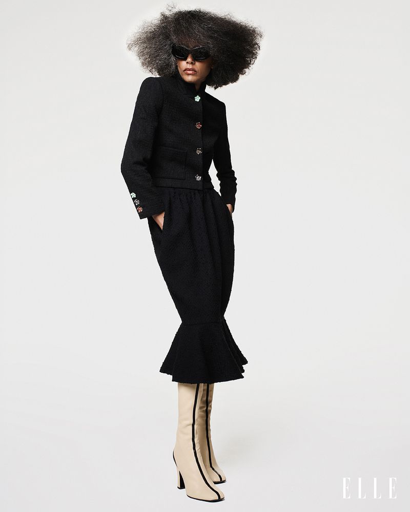 Model Stephanie Cavalli posing in a black skirt suit, sunglasses, and beige heeled boots against a white studio background.