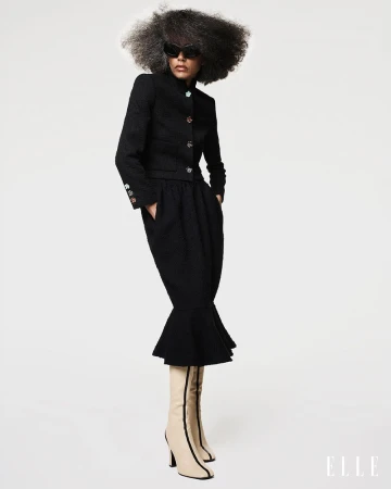 Model Stephanie Cavalli posing in a black skirt suit, sunglasses, and beige heeled boots against a white studio background.