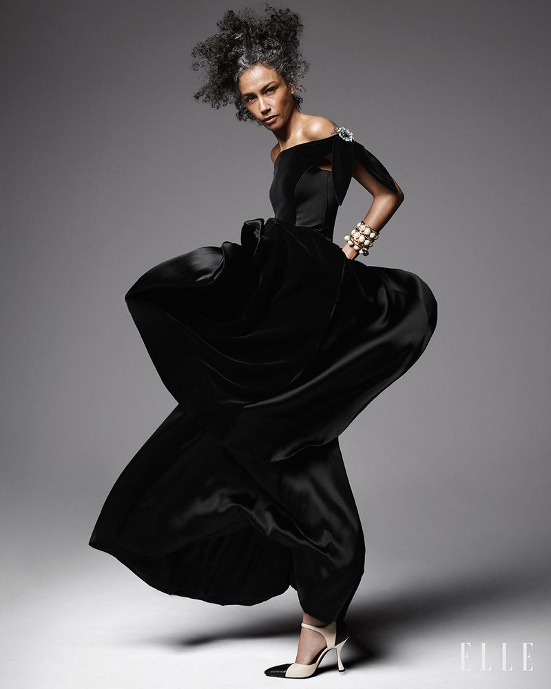 Model Stephanie Cavalli posing in a dynamic, sweeping black off-the-shoulder gown and two-tone heels against a grey studio background.