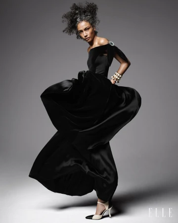 Model Stephanie Cavalli posing in a dynamic, sweeping black off-the-shoulder gown and two-tone heels against a grey studio background.