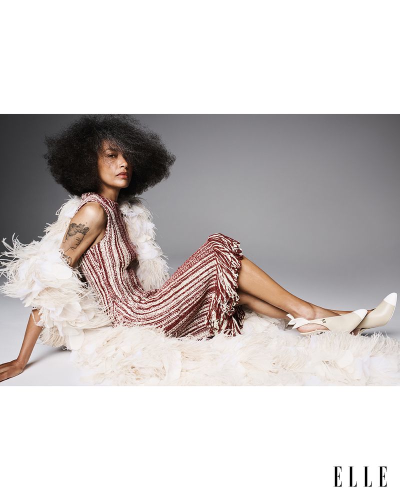 Stephanie Cavalli reclining in a burgundy striped fringe dress and voluminous white feather wrap on a grey studio backdrop.