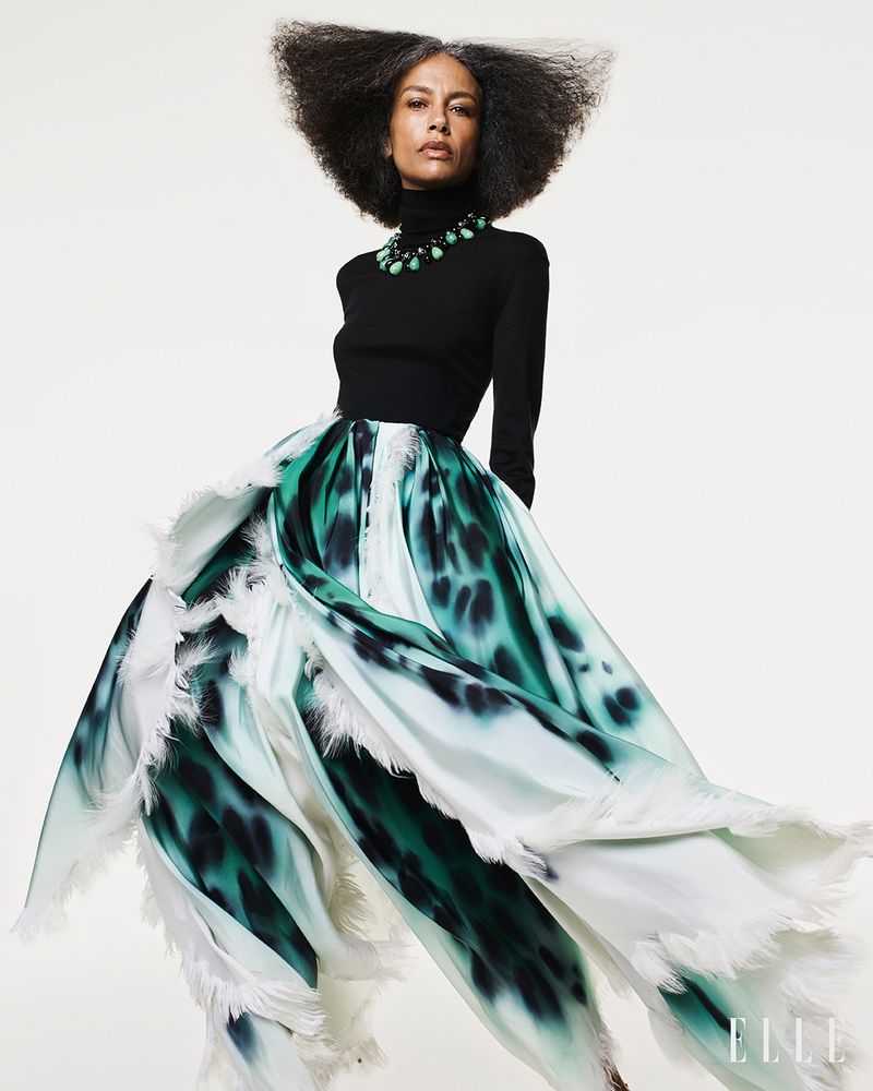 Model with voluminous dark hair wearing a black turtleneck and full green and white patterned skirt against white.