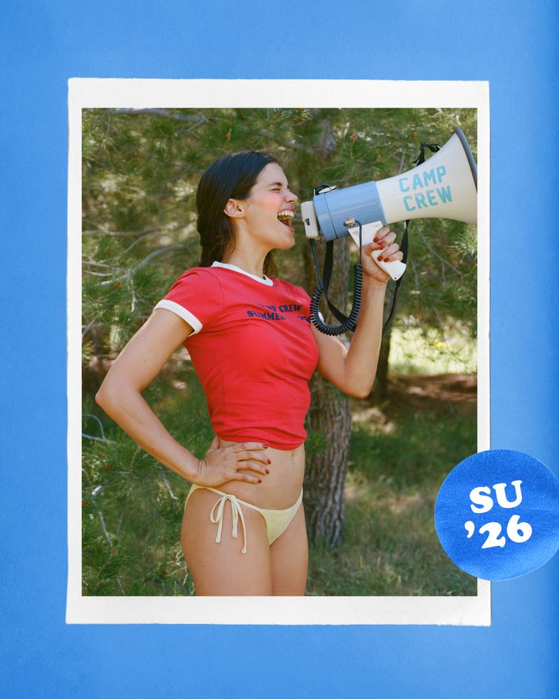 Sara Sampaio shouting into a blue and white megaphone wearing a red ringer t-shirt and bikini bottoms in a pine forest setting.