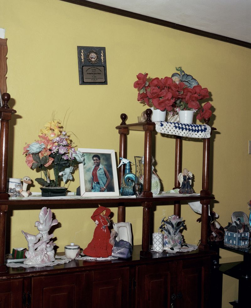 Home interior featuring a wooden shelf displaying assorted figurines, artificial flower arrangements, and a framed portrait on a yellow wall.