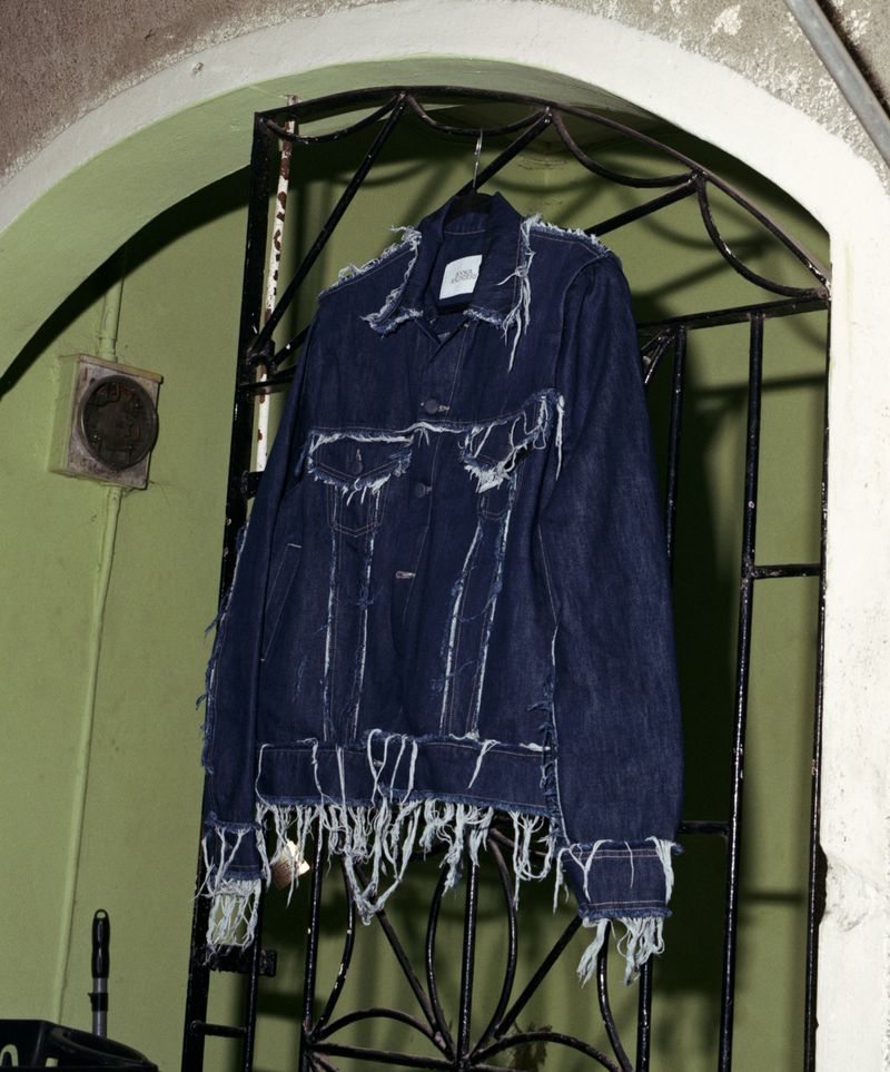 Hanging distressed denim jacket with fringed details, suspended from a metal gate with a green wall in the background.