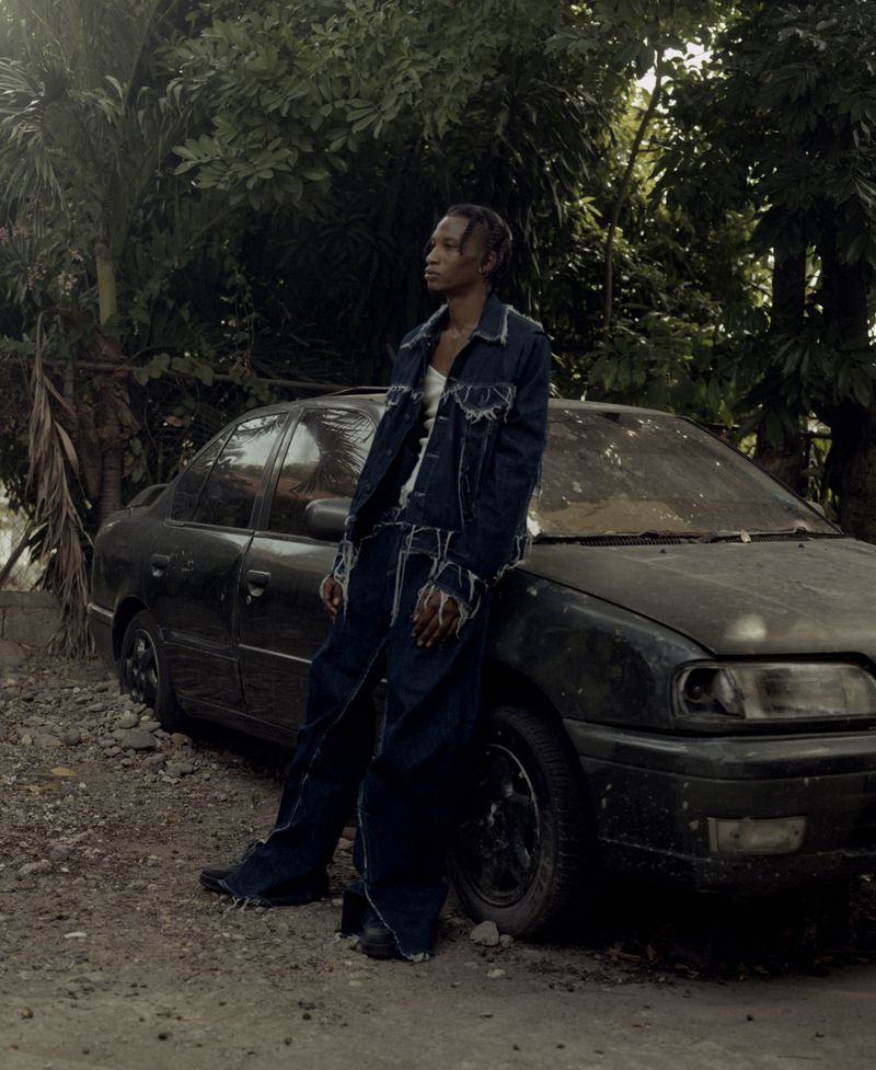 Young adult in a denim outfit leans against a weathered, dark car amidst lush foliage, styled in an outdoor, fashion editorial setting.
