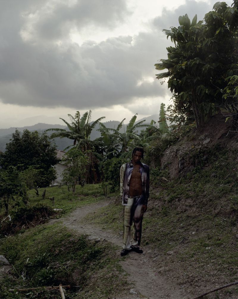 A full shot of a Black adult male standing on a dirt path in a rural area surrounded by tropical plants under a cloudy sky.