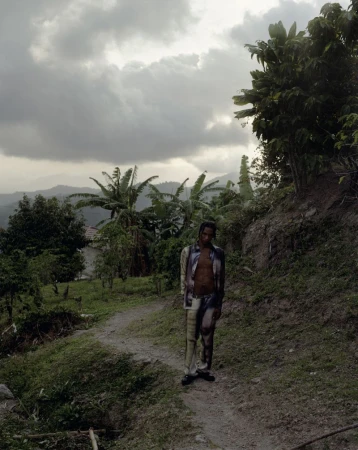 A full shot of a Black adult male standing on a dirt path in a rural area surrounded by tropical plants under a cloudy sky.