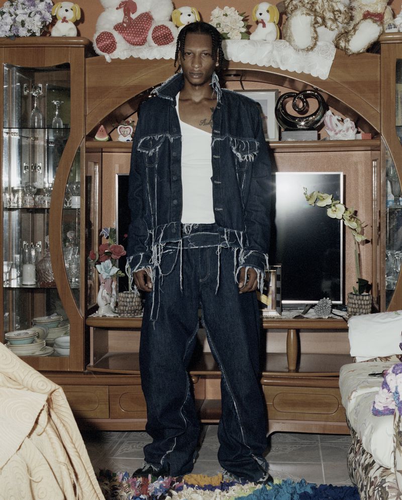 Full-length studio shot of an adult male wearing a dark denim outfit with white trim standing in an interior living room setting.