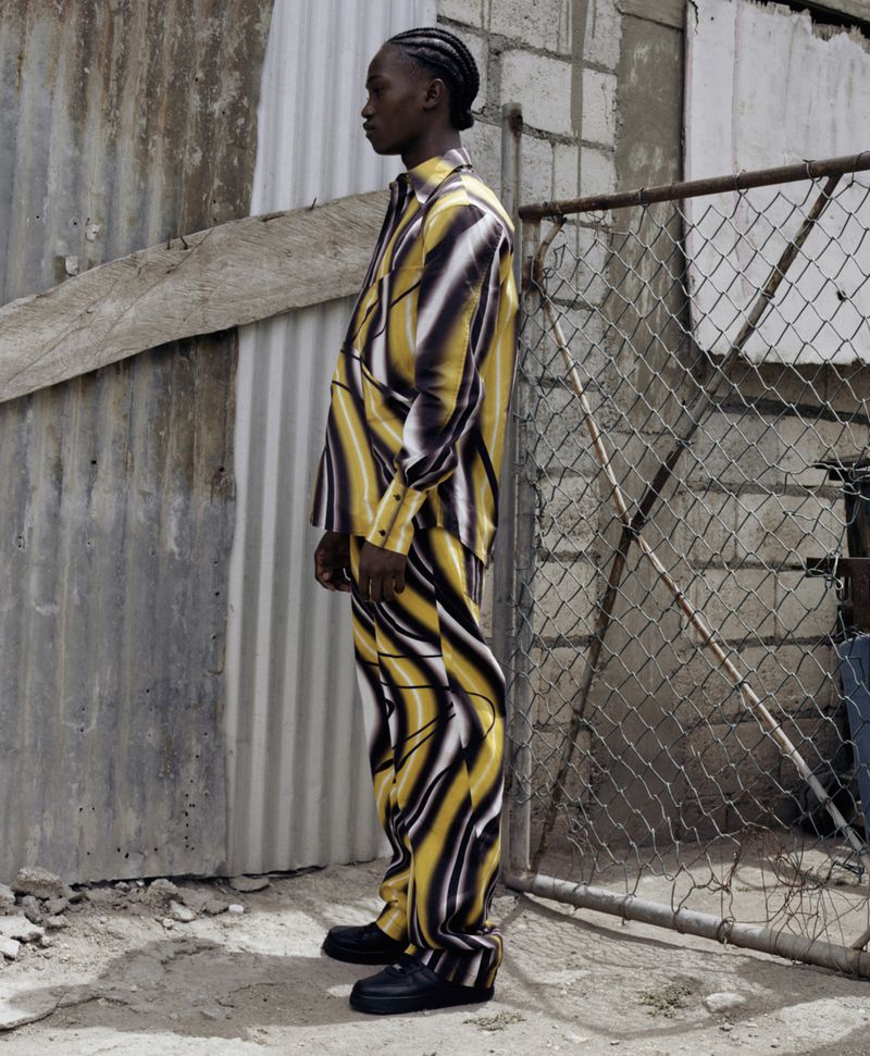 Adult male in patterned yellow and black suit stands in profile near metal fence with concrete and metal background in outdoor setting.
