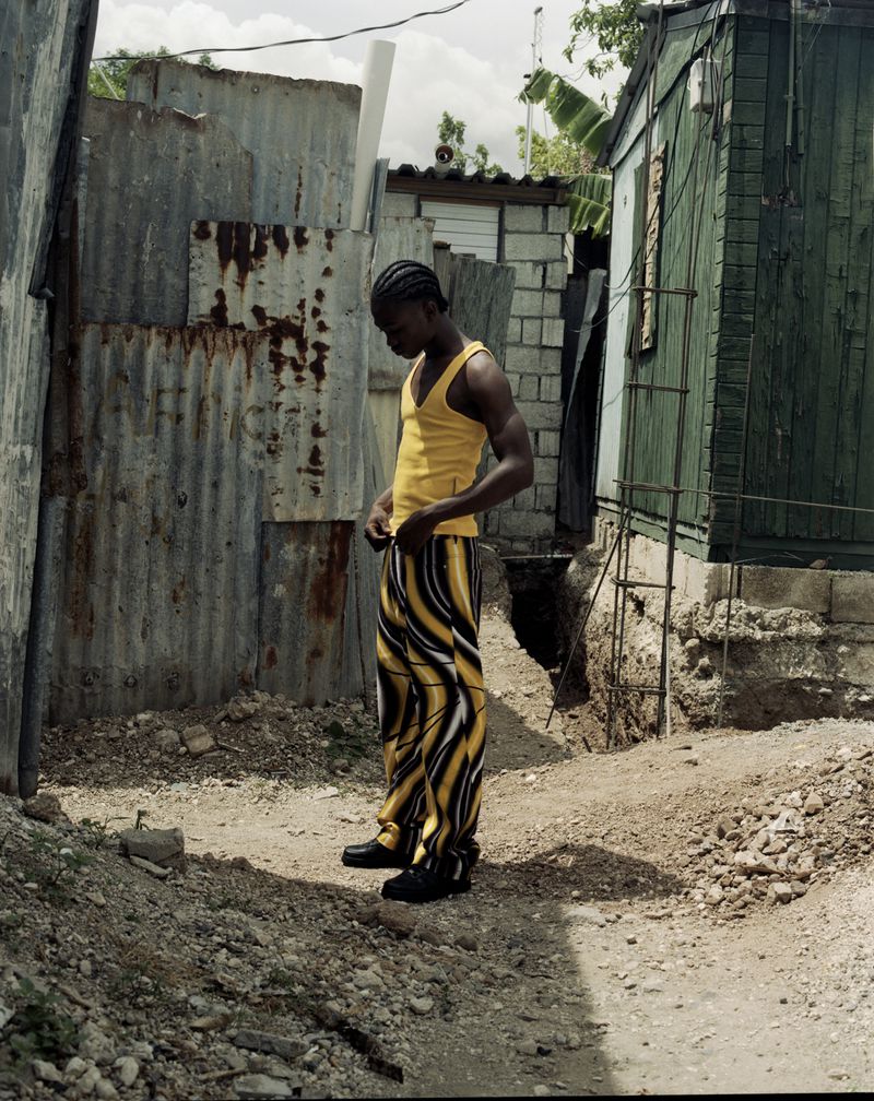 Man in yellow tank top and patterned pants stands outdoors near corrugated metal and building, bathed in natural light. 