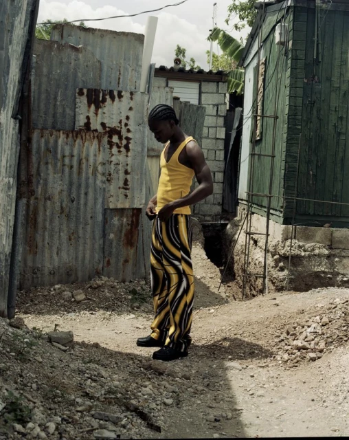 Man in yellow tank top and patterned pants stands outdoors near corrugated metal and building, bathed in natural light. 