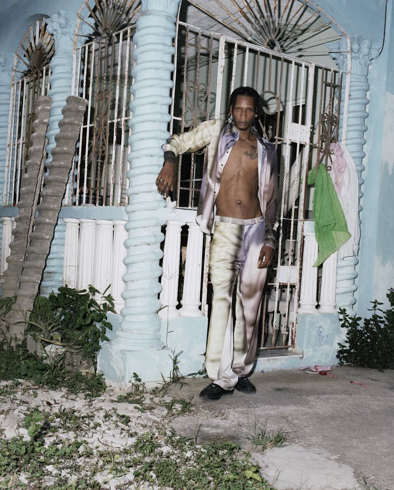 An adult male poses outdoors against a light-blue building in a patterned, matching set of an unbuttoned jacket and pants in a portrait shot.