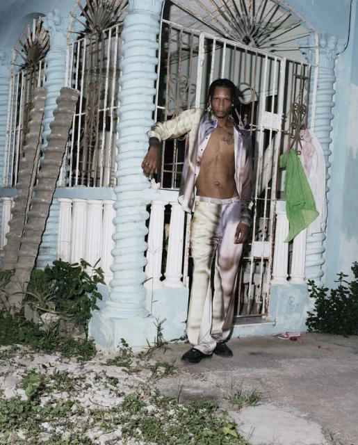 An adult male poses outdoors against a light-blue building in a patterned, matching set of an unbuttoned jacket and pants in a portrait shot.