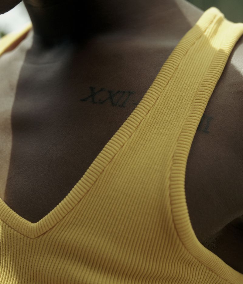 Close-up on a person with dark skin wearing a mustard yellow tank top and displaying Roman numeral tattoos on their upper chest.