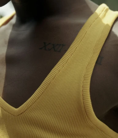 Close-up on a person with dark skin wearing a mustard yellow tank top and displaying Roman numeral tattoos on their upper chest.