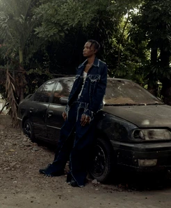 Man wearing a dark denim jacket and trousers leaning against a dark car outdoors surrounded by lush green tropical foliage.
