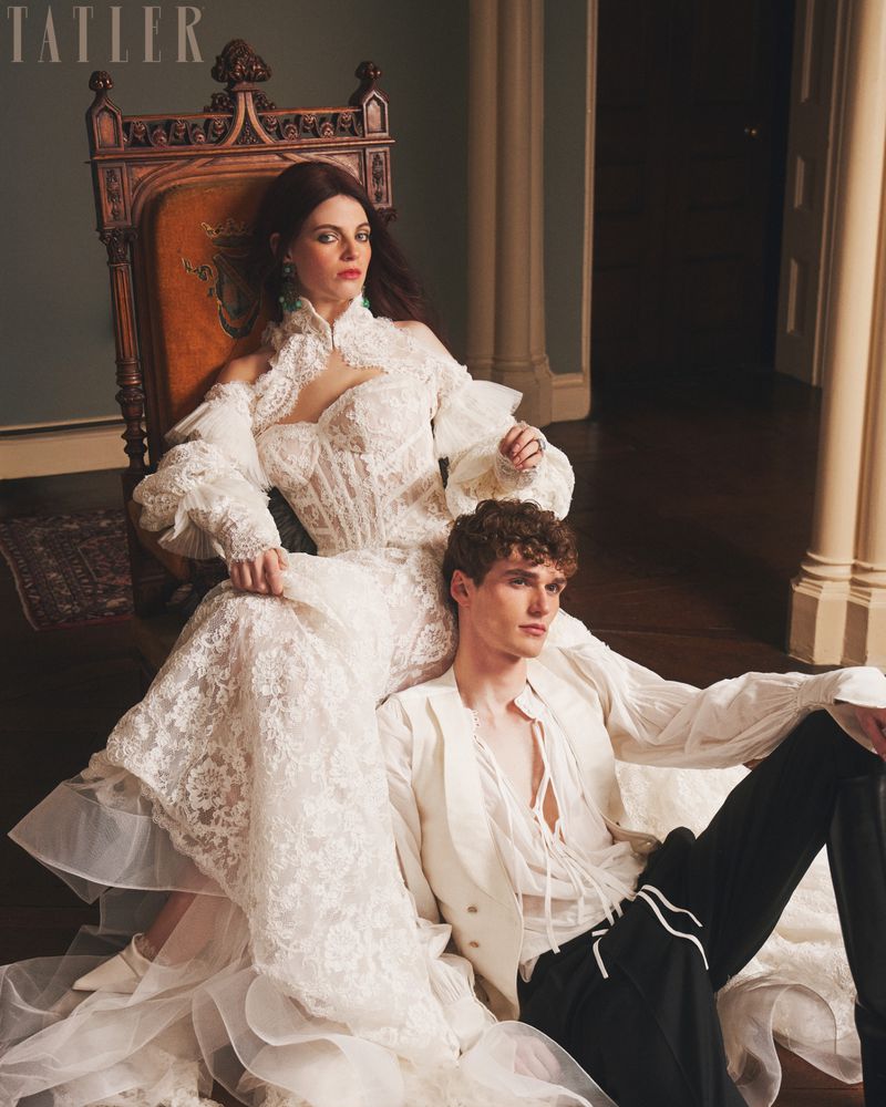 Fashion editorial shot of a woman in a white lace wedding dress sitting in an antique chair with a man in a white shirt next to her.