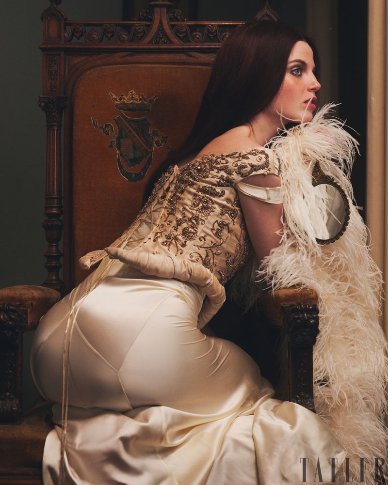 Brunette woman in an off-the-shoulder satin gown seated on an ornate chair, looking to the side with feathers and dark tones.