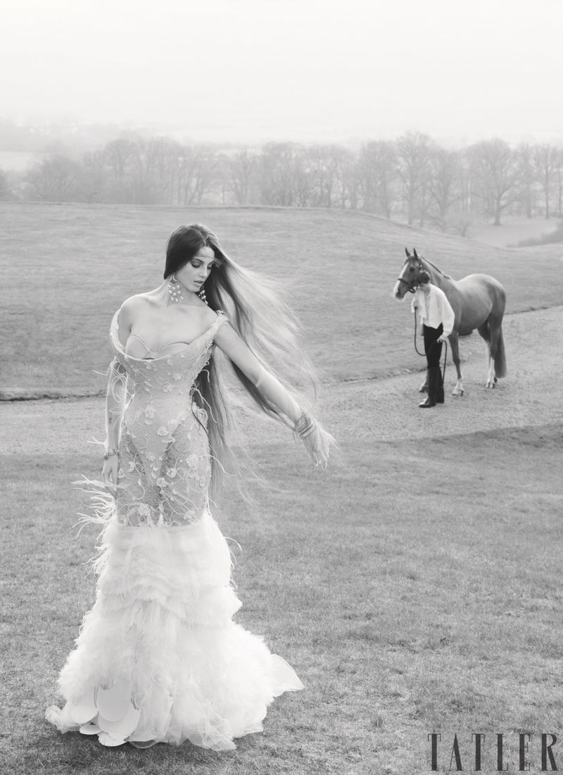 Model posing in an ornate feathered gown in a meadow with a horse and handler in the background.