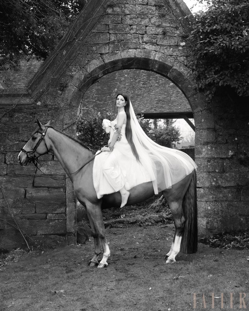 A woman in a wedding dress rides a brown horse through an arched stone gate, captured in a black and white editorial style photograph.