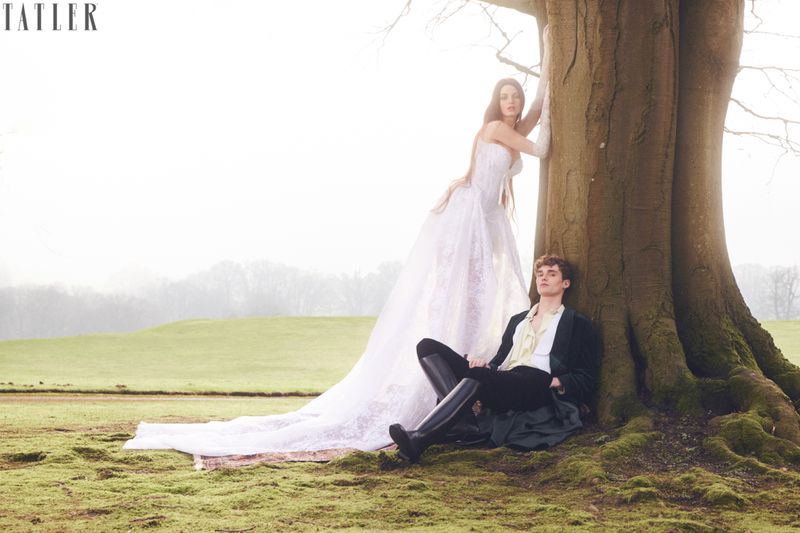 A couple poses at the base of a large tree in a field. The woman wears a white dress and the man is in a dark suit.