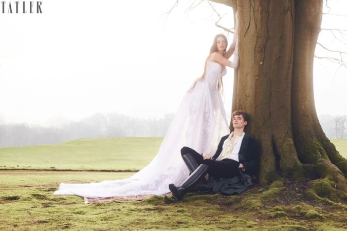 A couple poses at the base of a large tree in a field. The woman wears a white dress and the man is in a dark suit.