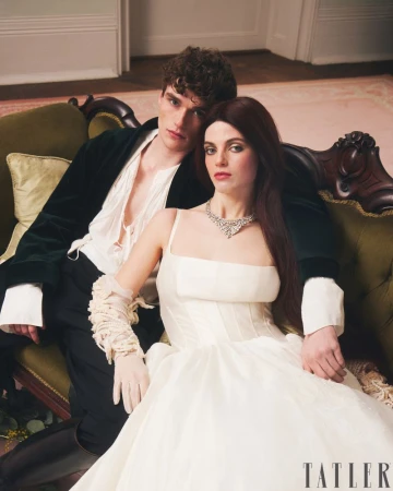 A stylish couple posing on an olive-green velvet couch. The woman wears a white gown and the man wears a black velvet jacket.