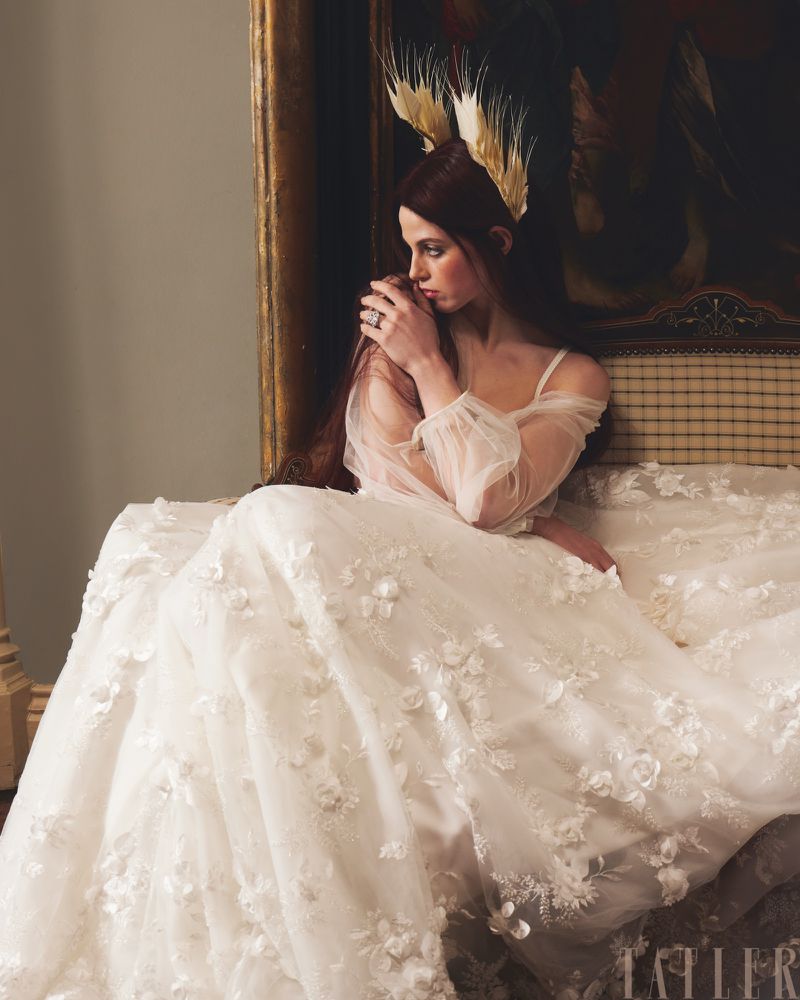 Elegant woman with long auburn hair wears a white, floral-embellished gown and feathered headpiece, seated against a draped backdrop.